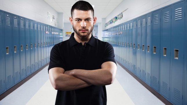 An angry looking guy with brown hair and brown eyes with his arms crossed standing in a hallway of high school lockers.
