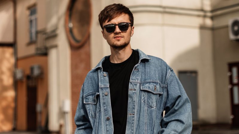 Attractive brown haired man in black sunglasses, a black sweatshirt, and a denim jacket standing in front of tan European style buildings.