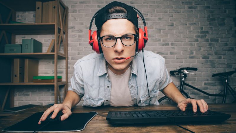 Angry young adult man with glasses and orange headphones glaring at a computer grabbing onto a computer mouse.