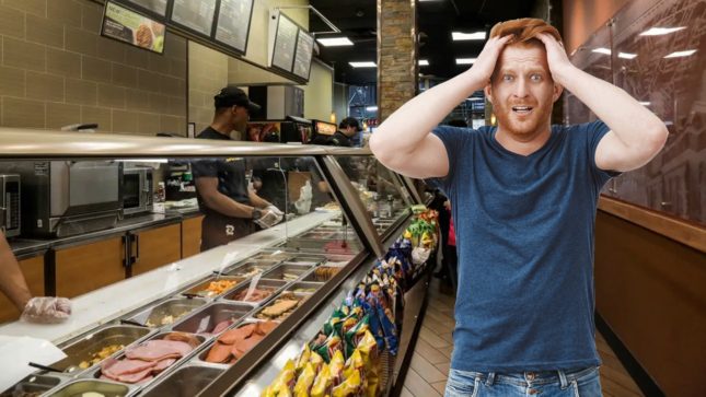 Red haired man in a blue shirt in line at subway looking panicked with both of his hands on both side of his head.