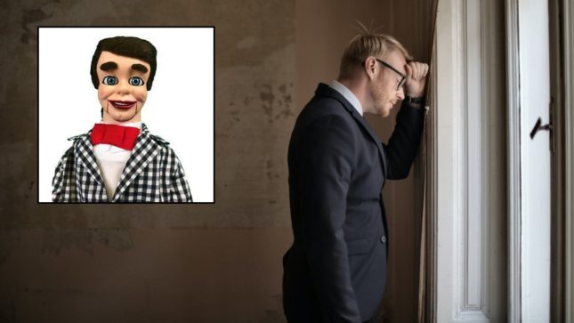 Blonde man in a suit leaning his head and fist against a wall looking sad, with a creepy looking, brunette ventriloquist dummy in a photo beside him wearing a red tux, white shirt, and checkered suit.