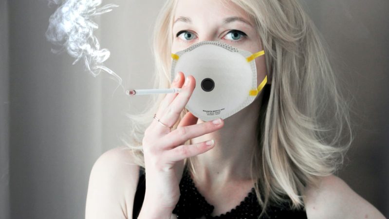 Blonde woman with green eyes smoking a cigarette through a face mask with a hole in it.