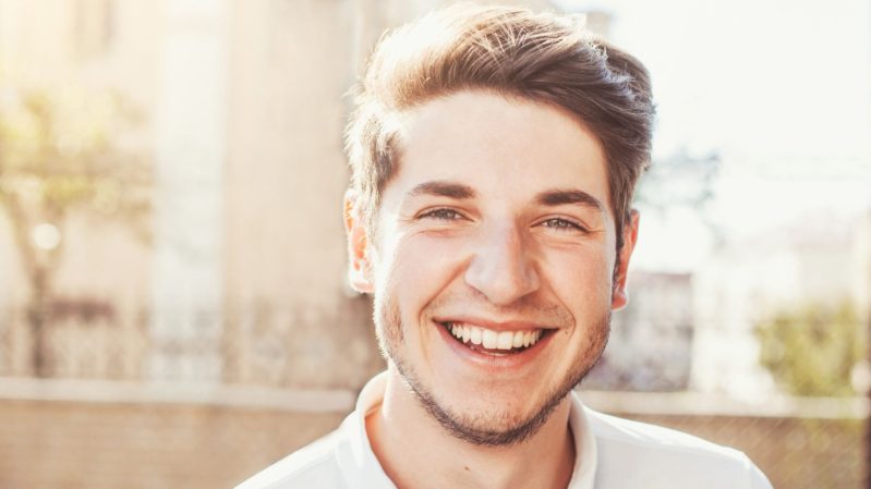 White boy with blonde hair and hazel eyes smiling with bright teeth and wearing a white, collared shirt against a beige background outside.