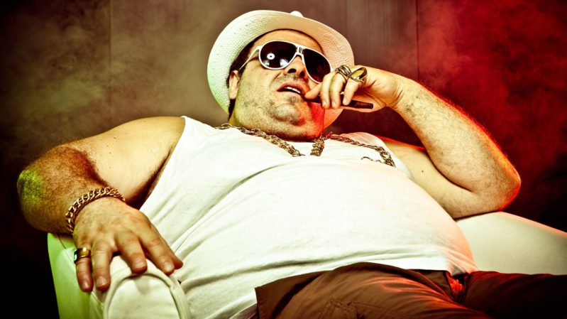 Italian mobster with cigar, heavy set in a white tanktop, white hat, sunglasses and brown pants sitting down with smoke in the background.