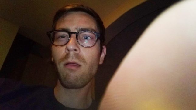 Brown haired, blue eyed pale man in glasses with a confused look on his face in a selfie wiht his hand half over the camera.