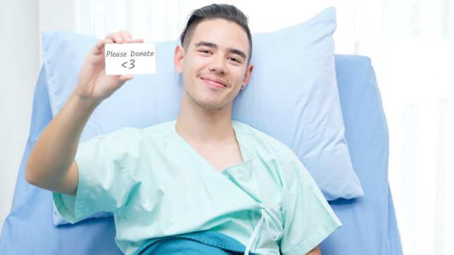 Man smirking in hospital bed holding up a small white card that says "Please Donate" with a heart emoticon.