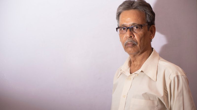Tan, south asian man in glasses with gray hair with a dry expression wearing a pale yellow collared shirt in dim lighting against a white, pinkish wall with brown eyes.