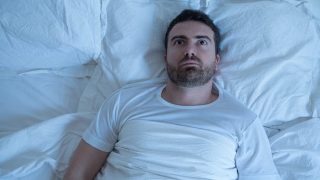 Pale man with dark brown hair and brown eyes wearing a white t-shirt, laying on white pillows and sheets. He looks like he can't sleep because he is stressed and anxious.