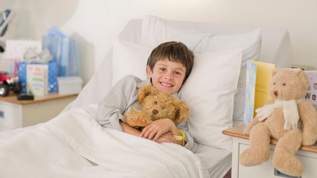 Little boy in hospital bed with brown hair and brown eyes, olive pale skin and gapped teeth, smiling and cuddling a teddy bear in his striped pajamas and is under the covers.