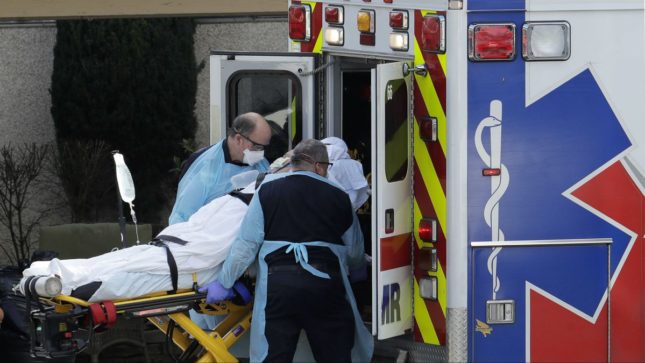 Body wrapped in a white blanket on a turnakit being rolled into an ambulance, cannot see the woman's face, two EMT's in EMT outfits.