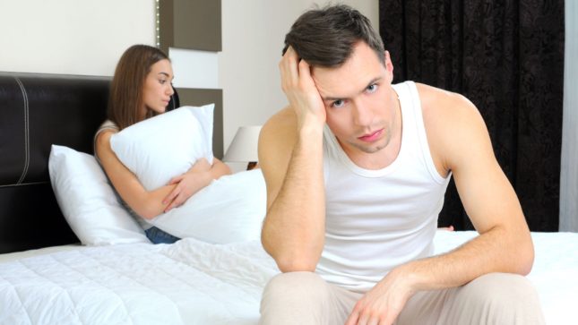 Man in bed looking at the camera angry and woman in bed holding a pillow. The man has his hand on his head and blue eyes, pale skin, and brown hair. The woman has brown skin, brown eyes, and brown hair.