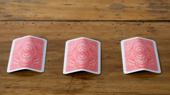 The back of three red cards with a white outline on a tan, wood table.