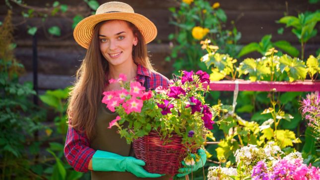 Woman with long, brown, straight hair and big, green eyes wearing a yellow hat is holding a plant with pinkish and purple flowers. The basket is brown. She is wearing a red and dark blue checkered shirt and bright green gloves. She is standing in front of a garden of flowers.