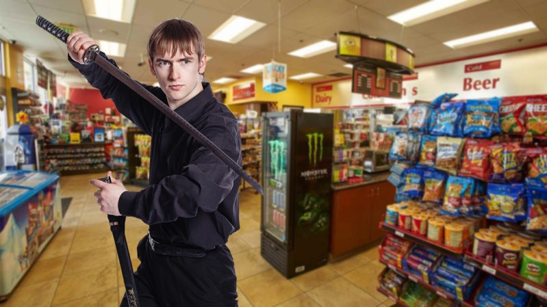 Dark blonde, blue eyed scrawny man in a black karate outfit with a sword standing in a gas station posed in a fighting position.