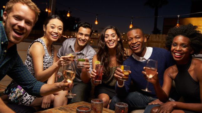 Friend group smiling with drinks outside at a roof top bar with hanging lights in the background.