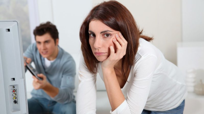 Man with brown hair and tan skin blue collar shirt long sleeves playing video games. Woman staring into the camera annoyed, looking fed up with it. She has brown hair with a red tint, olive-pale skin and brown eyes.