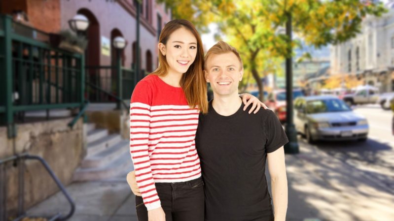 Tall, asian woman with light brown hair in a red and white striped shirt crouching next to a short, white, blonde man in a black shirt.