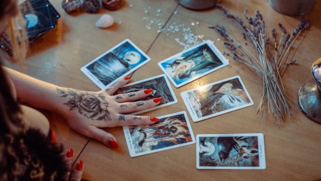 Hand with red nails hovering above six tarot cards on a table in dim light.