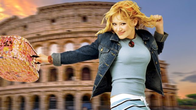 Hilary Duff as Lizzie McGuire at age 15 spinning in front of the colosseum in a blue shirt, denim jacket, and towl blue and black striped shirt holding an orange hand bag.
