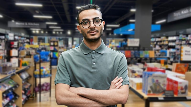 Man with black hair, dark brown eyes, light brown skin, black beard and black glasses standing in a mall. He is wearign a muted green button-up shirt buttoned to the top button, with white buttons. He has his arms crossed and looks annoyed.