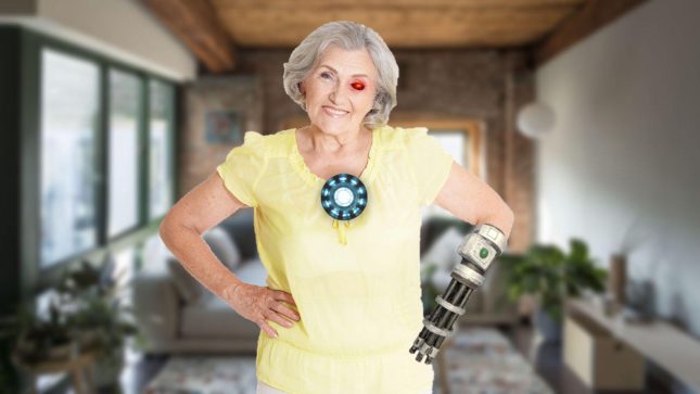 Grandmother in a yellow shirt with short grey hair smiling. She has a laser red eye and is wearing a pale yellow shirt. She has a grey and blue button in the middle of her chest with a white center. Both of her hands are on her hips and she has a robotic right forearm. She is behind a conventional living room with windows, a white wall, and a tan couch with hard-wood floors.