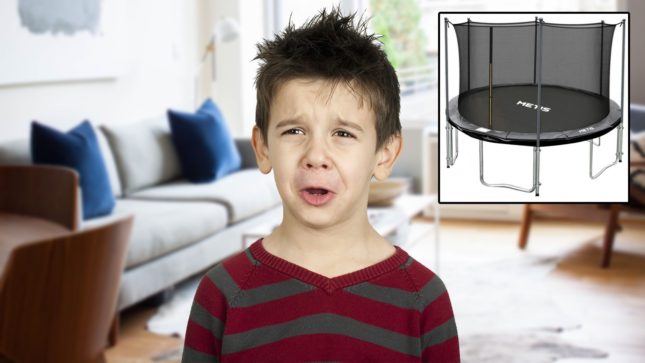 Kid with olive skin and brown hair looking like he's whining and crying. He is in a living room with a beige carpet and gray couch with blue pillows and large windows. Beside him in a square photoshopped in there is a trampoline. He is wearing a dark red and dark gray striped shirt.