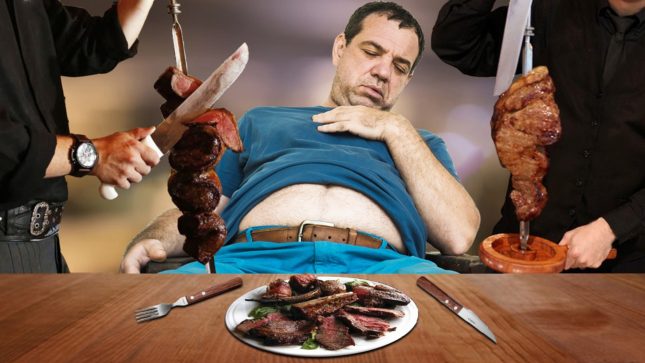 Man with a big belly and his blue t-shirt rolled up with his eyes closed surrounded by bloody knives with a plate of meat in front of him on a wood table with a fork and knife on either side of him.