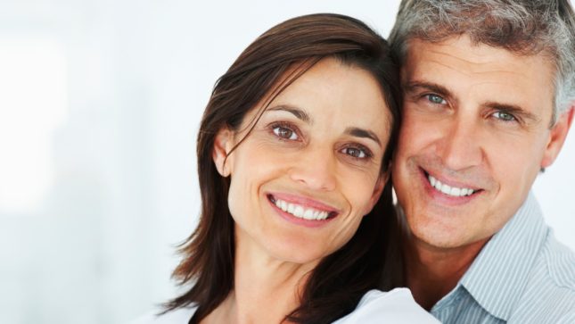 Woman with black hair and blue eyes and pale skin smiling next to a man with pale skin, blue eyes, and gray hair smiling. Both are middle aged and are standing in front of a white background.
