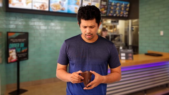 Tan man with black eyes in a dark blue shirt looking disappointed into an empty wallet. Standing in front of a fast food restaurant in a food court that has a menu board and light green background and a counter.
