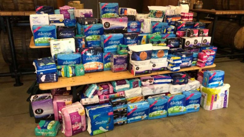 Picture of various tampons and pads stacked on each other on a light wood table. The floor is a tiled gray-green and there are two dark wood tables beside it.