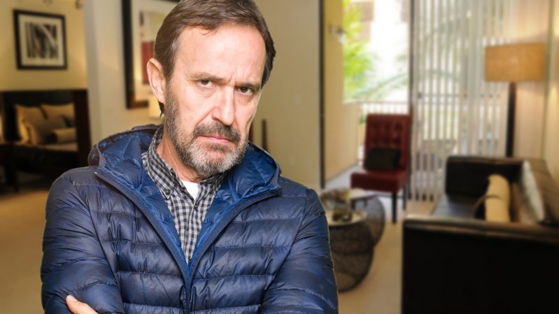 Angry, ashy-white middle-aged man with brown hair and a mustache against a living room background. He has brown eyes, and is wearing a padded navy blue jacket.