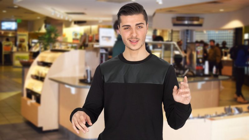 Drunk guy with black hair and brown eyes and an out-of-it expression, wearing a long sleeve black shirt, standing in front of a dining hall.