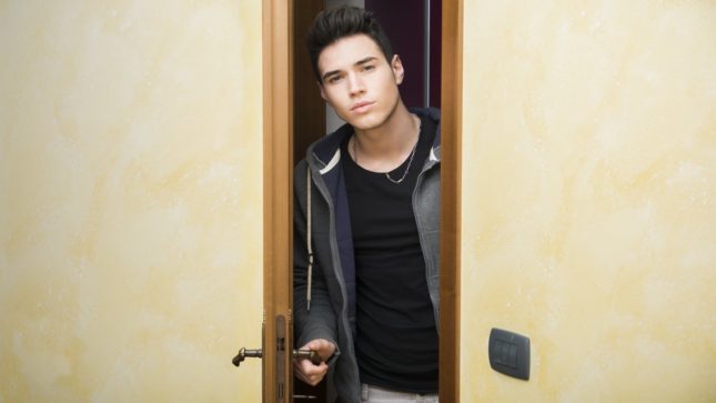 College aged guy with pale skin, black hair, and brown hair poking his head out of the door.