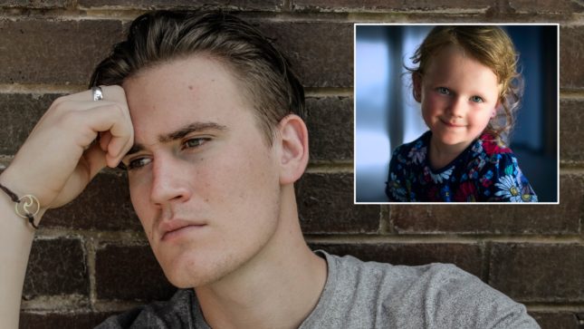 White man with wide jaw and greenish brown eyes and light brown hair resting his fist on his forehead looking distressed. In the corner is a picture of a young, elementary school aged brown haired pale girl with blue eyes in a blue shirt. He is against a brick wall with a light green shirt on wearing a bracelet and a silver ring.