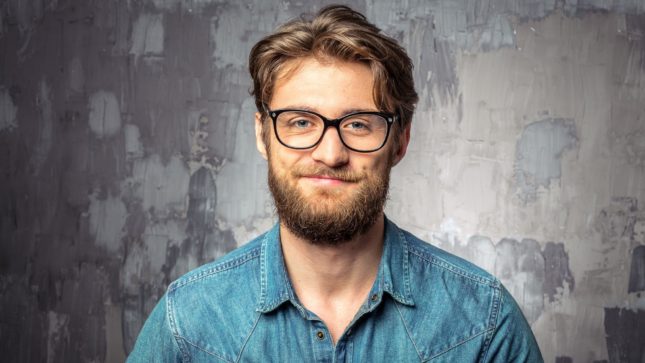 Pale, white man with small brown eyes and big framed glasses against a grey wall. He has a light brown, full beard and medium-length light brown hair. He's grinning. Wearing a collared blue shirt.