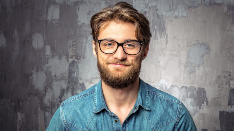 Pale, white man with small brown eyes and big framed glasses against a grey wall. He has a light brown, full beard and medium-length light brown hair. He's grinning. Wearing a collared blue shirt.