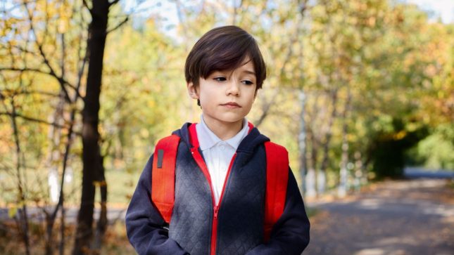 Brown haired white boy about 10 years old looking down at the ground with a sad expression. Against an autum trees background, with green and yellow leaves. He is standing on the pavement. He is wearing a blue button up shirt, a navy sweatshirt with a red zipper, and has a red backpack on.