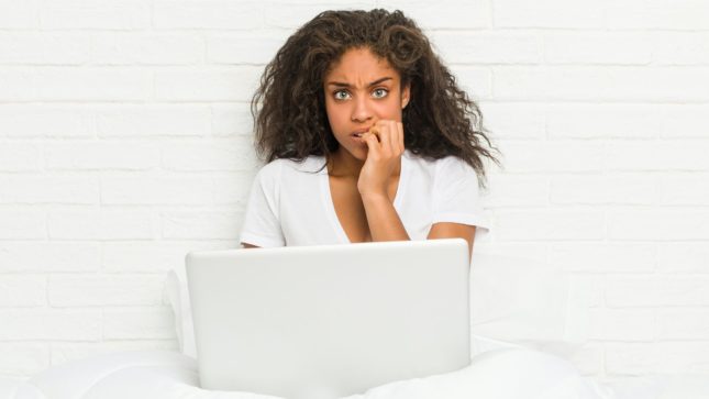 Woman with frizzy black hair, big green eyes, and blakc skin biting her nails, looking frightful. She is sitting under the covers on her bed with her white laptop in front of her as if video chatting, wearing a white t-shirt and sitting against a white brick wall.