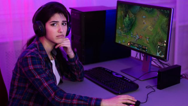 Girl with tan skin, short black hair, and brown eyes looking annoyed as she stares at her computer screen with a video game on it. Purple background, white curtains, grey desk. She's wearing a green, black, and red flannel and a white t-shirt, and is resting her chin on her fist.