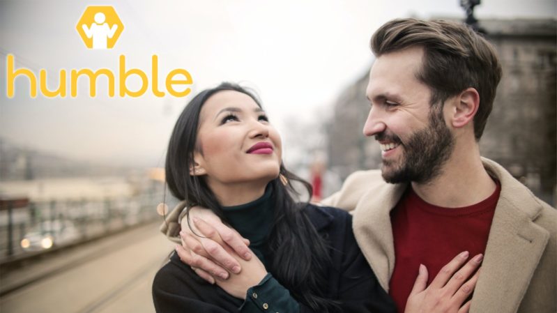 Smiling asian woman with red lips and straight black hair wearing a black turtleneck with her hand on the chest of a smiling ashy white man with brown hair and a brown beard, a red shirt, and a tan jacket, both standing on a gray background road. There's a "Humble" logo with a shrugging yellow and white figure in it.