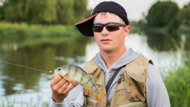 Fuckboy with fish and narrow rectangle black sunglasses, holding a fish. White, blonde, standing in front of a pond with a tan fisher's vest and a blue-ish-gray hoodie.