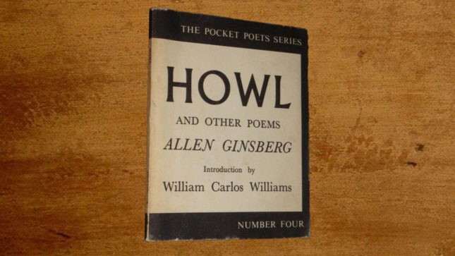 A white-tan book that says "HOWL" in black, large letters, and then Allen Ginsberg in black letters beneath it. It is on a light brown wood table. The book has a black line on the top and bottom.
