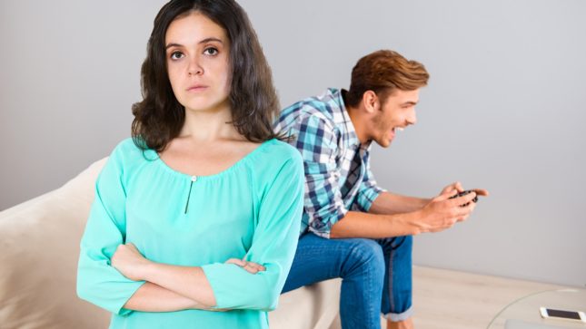 Sad woman with pallid olive skin and arms crossed with brown hair and brown eyes. She is wearing a turquoise quarter-sleeve shirt. The man next to her is on the couch playing video games. He is white and has light brown hair. He is playing video games on a tan couch and there is a gray wall behind them.