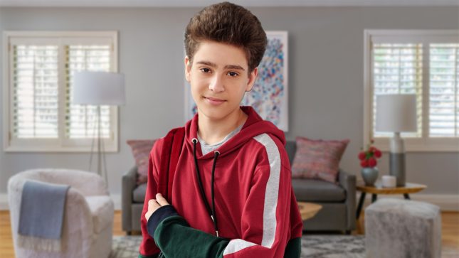 Tan boy in early teens and brown eyes standing with his arms crossed. He's in a gray living room with white windows. There is a gray couch with a blue blanket, and four white windows with crown molding and a crossed pattern on them. He has light brown hair that is fluffy and thick. He's wearing a red sweatshirt with black sleeves and his arm is crossed. The floor is white.