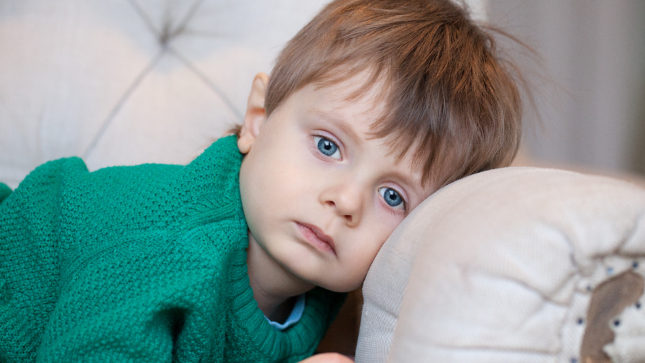 White blonde toddler boy with sad, blue eyes resting his head against a white couch. He is wearing a green long-sleeve shirt. He looks upset. There is a greyish white blurred wall behind him.