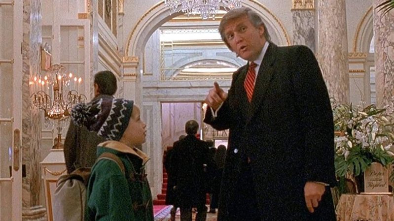 Fat orange man Donald Trump in a black suit and red tie pointing towards the camera. Standing next to him is shorter young boy Kevin McAllister looking up at him in a green jacket and a blue hat that has a pom-pom on top. They are in the St. Regis Hotel which is fancy and white with nice arches.
