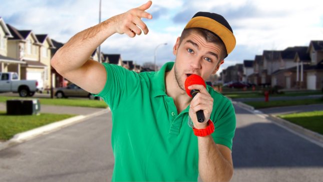 Olive skinned white boy with brown hair and brown eyes flashing a peace sign. He is wearing a black and brown brim hat. He is holding a red and black microphone to his lips. He is wearing a green collared shirt. He is standing in a suburban neighborhood on a sunny day.