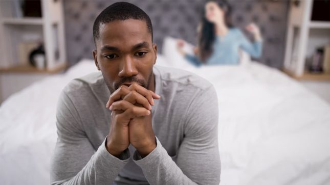 Black man in a gray shirt with his fingers interlocked, looking upset and contemplatitve. He is sitting on a bed with a white quilt and a grey headboard. There is a smiling white girl with long brown hair who is smiling and stretching, wearing a blue shirt.