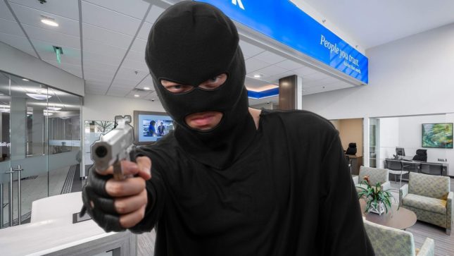 White man with blue eyes in a black ski mask pointing gun to the camera. He is standing in a bank that has white walls and a single blue wall near the gray, tile-lined ceiling.