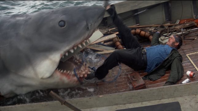 Jaws, a large shark, with his mouth on a small boat as a white man with black pants and a blue shirt screaming and sliding into his mouth.